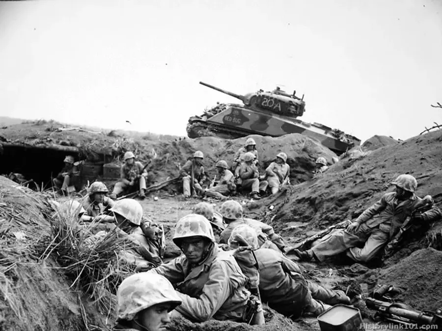 Marines of the 24th Marine Regiment on Iwo Jima, with a tank on the ridge above them.